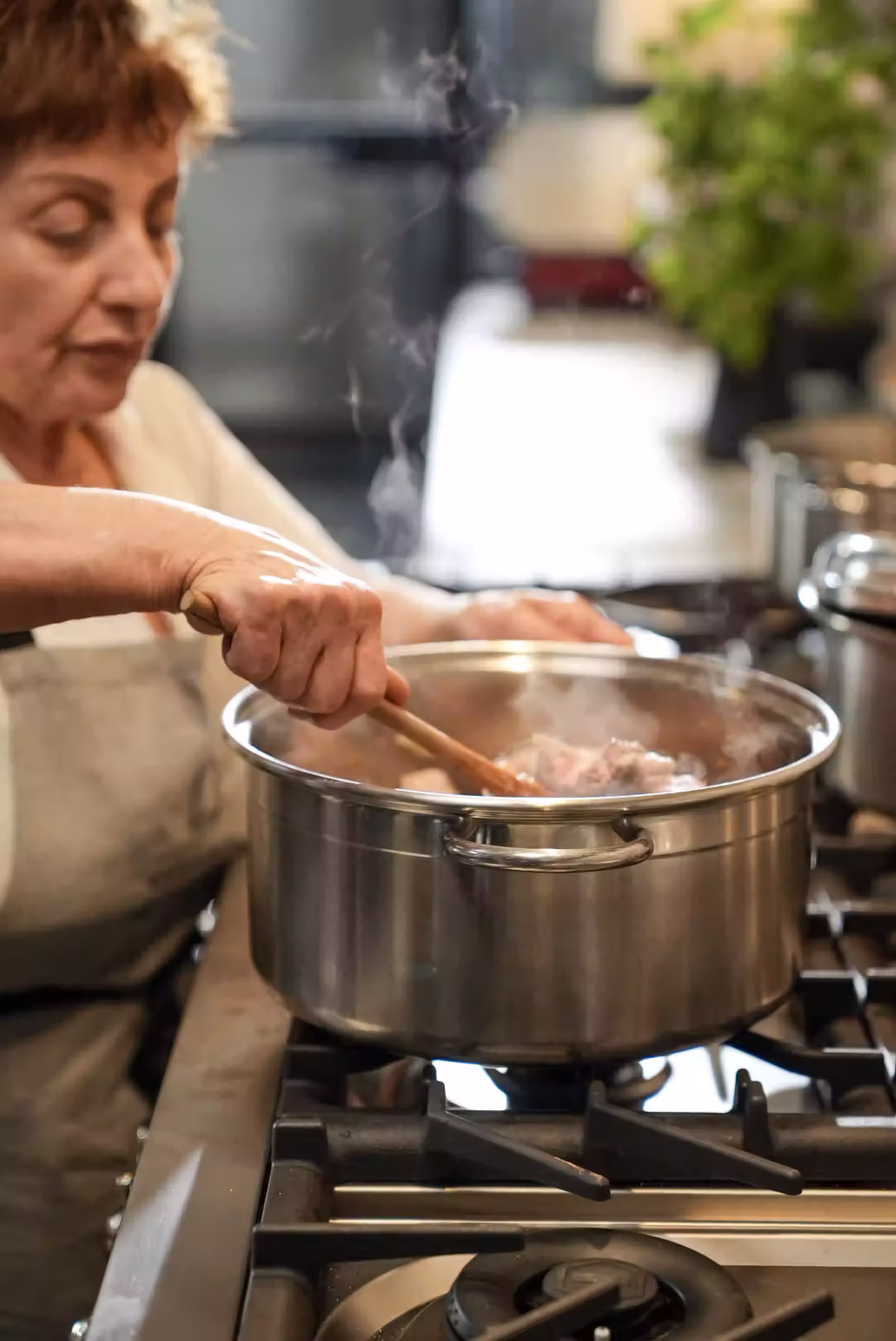 En äldre kvinna rör om i en ångande kastrull av rostfritt stål på spisen, iförd ljus förkläde, med mjuk köksbakgrund och grönska.