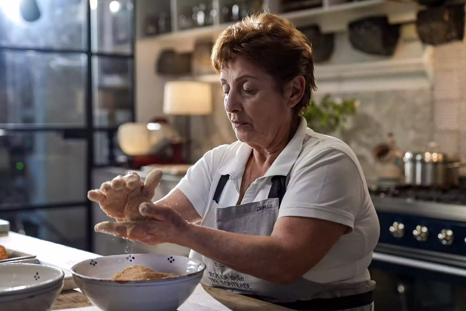 En äldre kvinna panerar mat över en skål, iförd förkläde och vit pikétröja, med köksredskap och spis i bakgrunden.