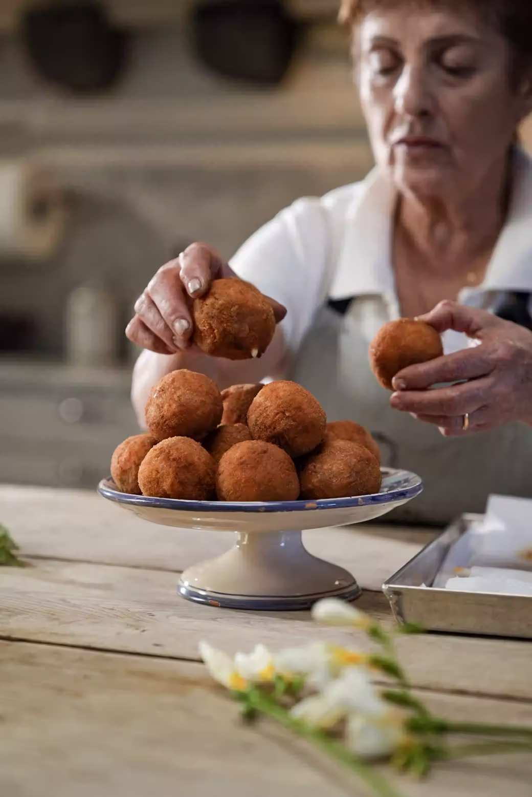 En äldre kvinna placerar en gyllenbrun friterad boll på ett tårtfat med lika bollar, vid träbord med blommor i rustikt kök.