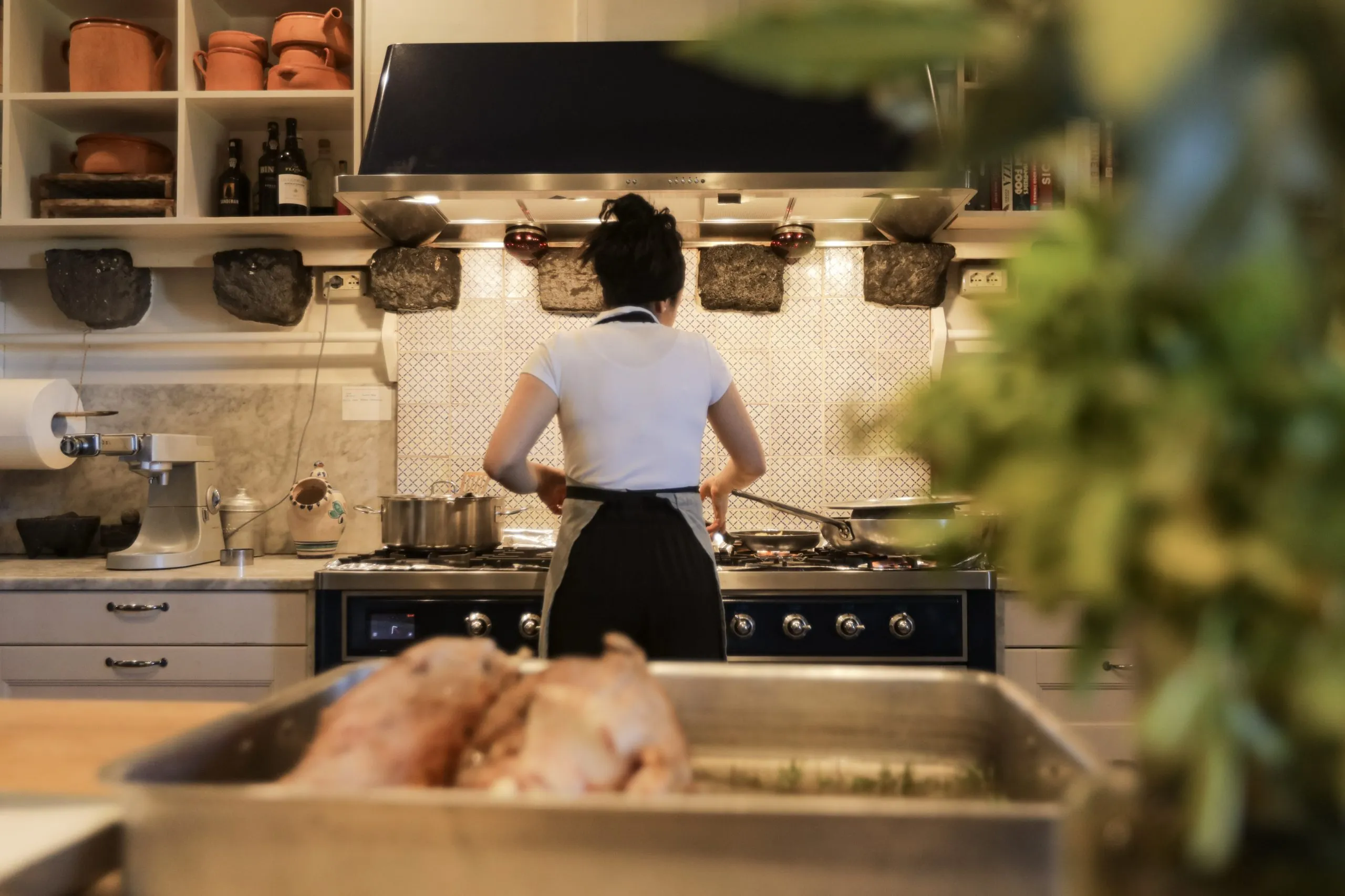 En kvinna i förkläde lagar mat vid spisen i ett ombonat kök, med hyllor för grytor och flaskor samt en bricka med fågel.