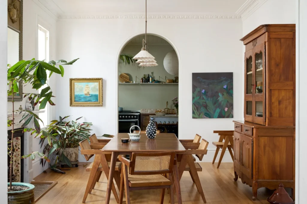 Ljust matrum med bord i trä, rottingstolar, stora växter och skänk; genom valv syns kök med hyllor och spis.