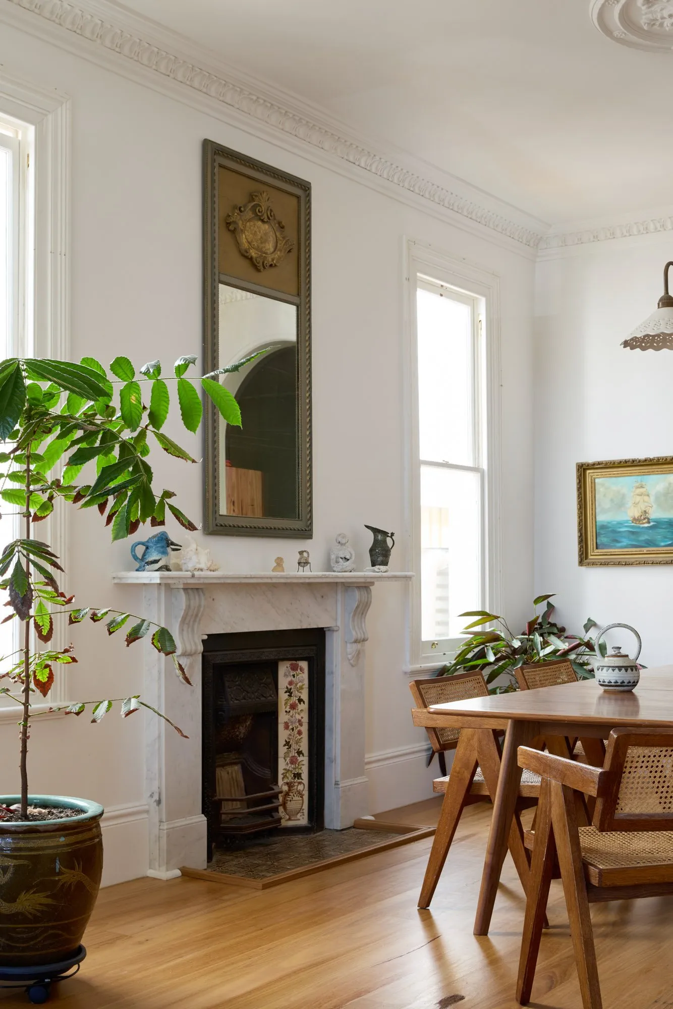 Ljust matrum med träbord och stolar, stor krukväxt, marmorspis med dekoration, höga fönster, spegel och tavla.