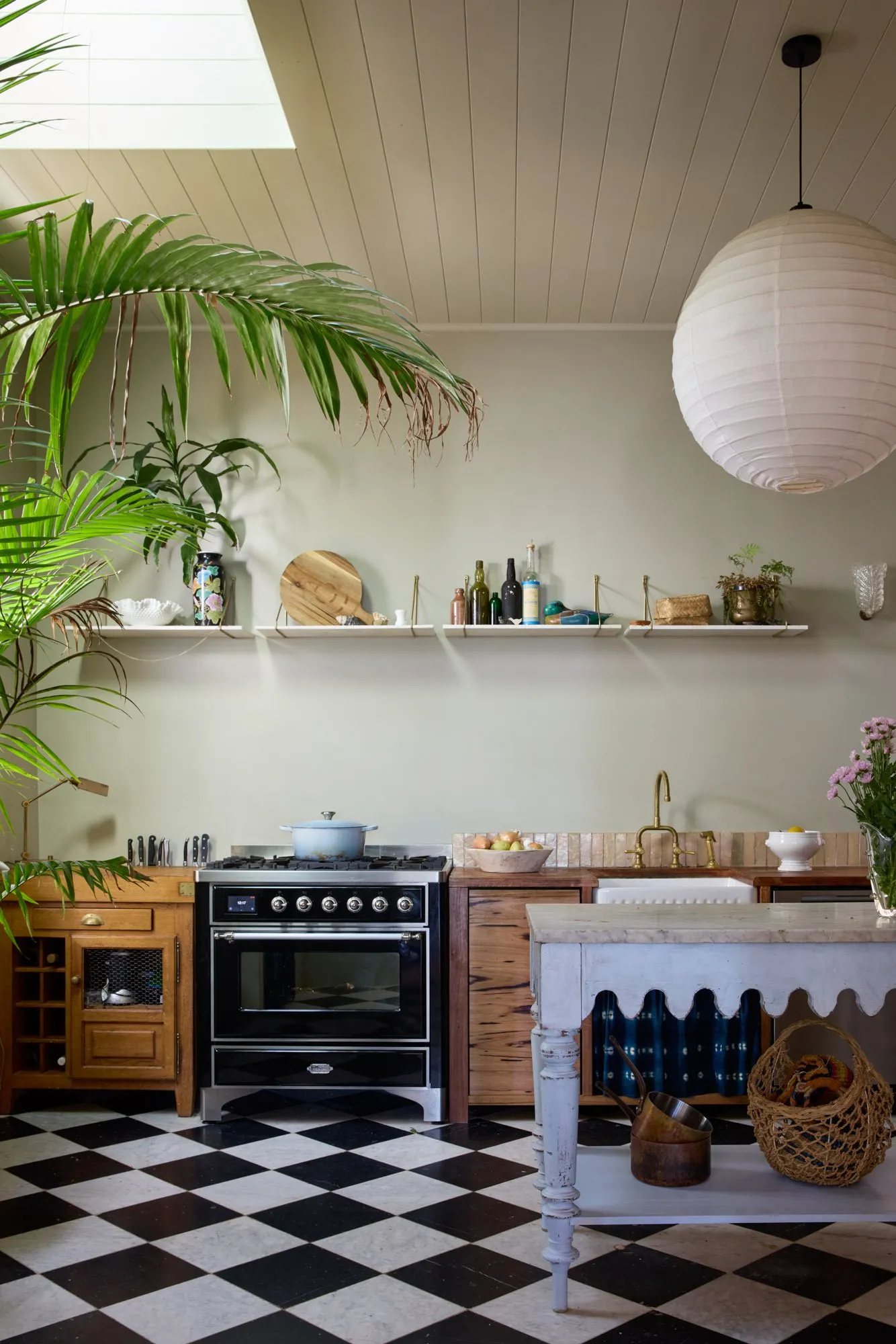 Kök med svart ugn, schackrutigt golv, träskåp, öppna hyllor med porslin och växter, stor palm och rund papperslampa.