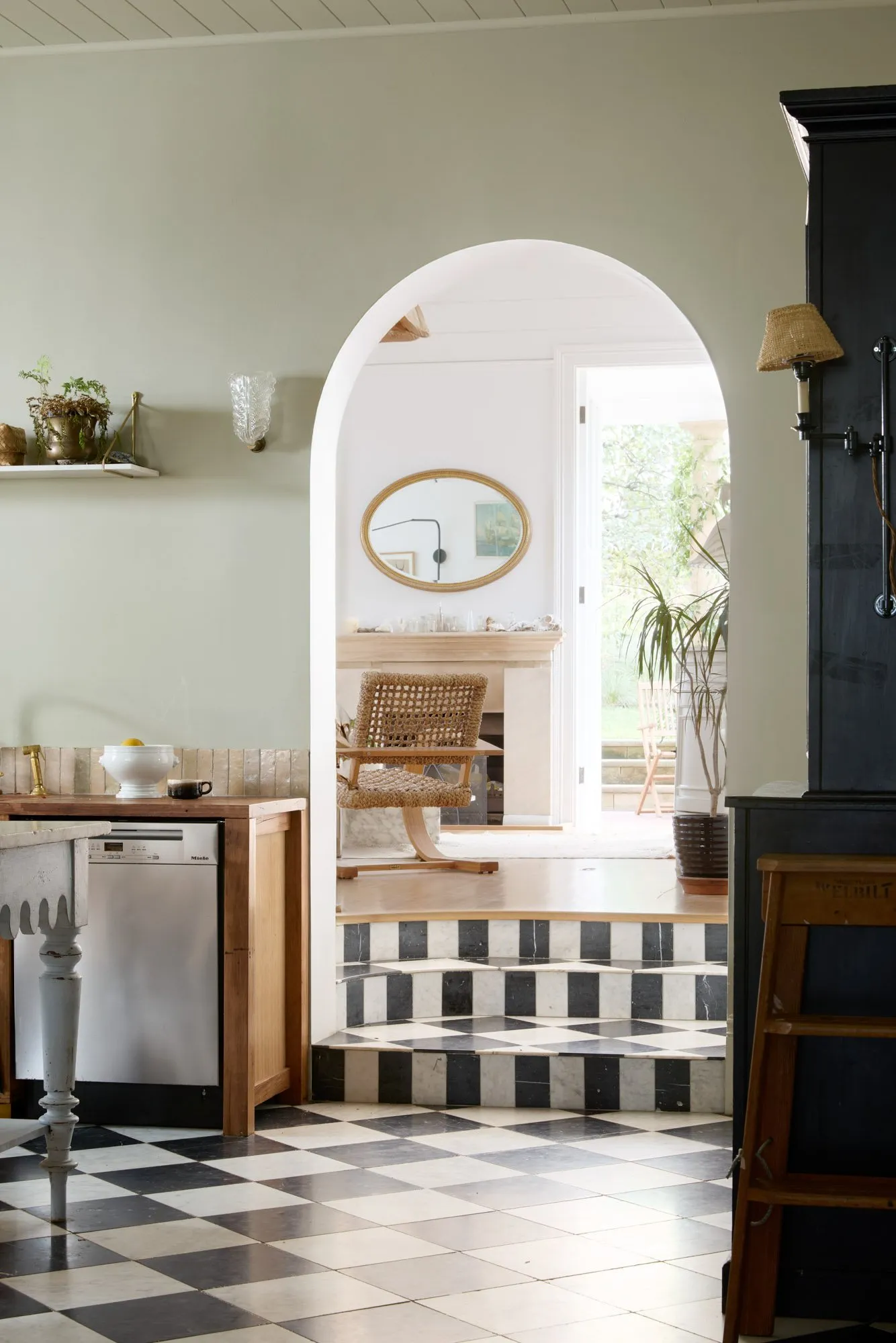 Ett kök med svartvitt rutigt golv, spis och diskho ansluter via valvbåge till ett ljust rum med spegel, gungstol och växter.