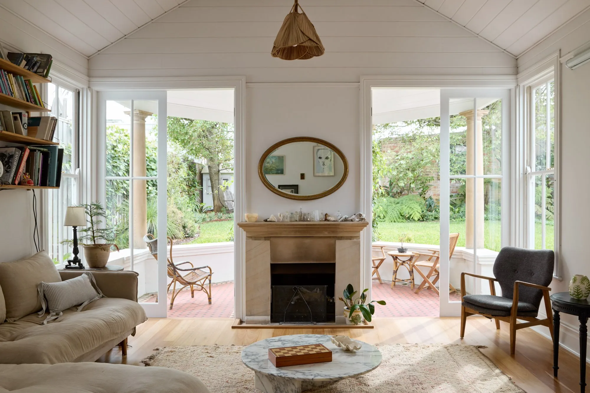 Ljust och ombonat vardagsrum med stora fönster, beige soffa, fåtölj, eldstad med runt spegelglas samt trä- och soffbord.