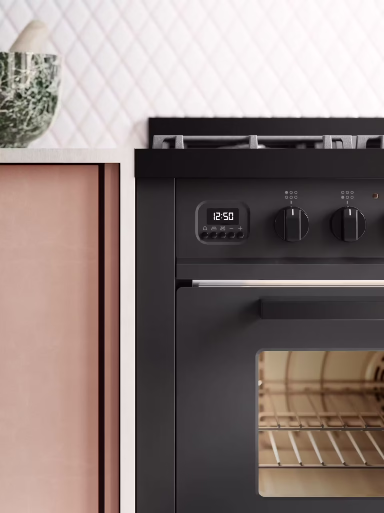 Detaljbild av svart Pro Line spis, 90 cm bred, intill rosa skåp; digital klocka visar 12:50 och mönstrat stänkskydd bakom.