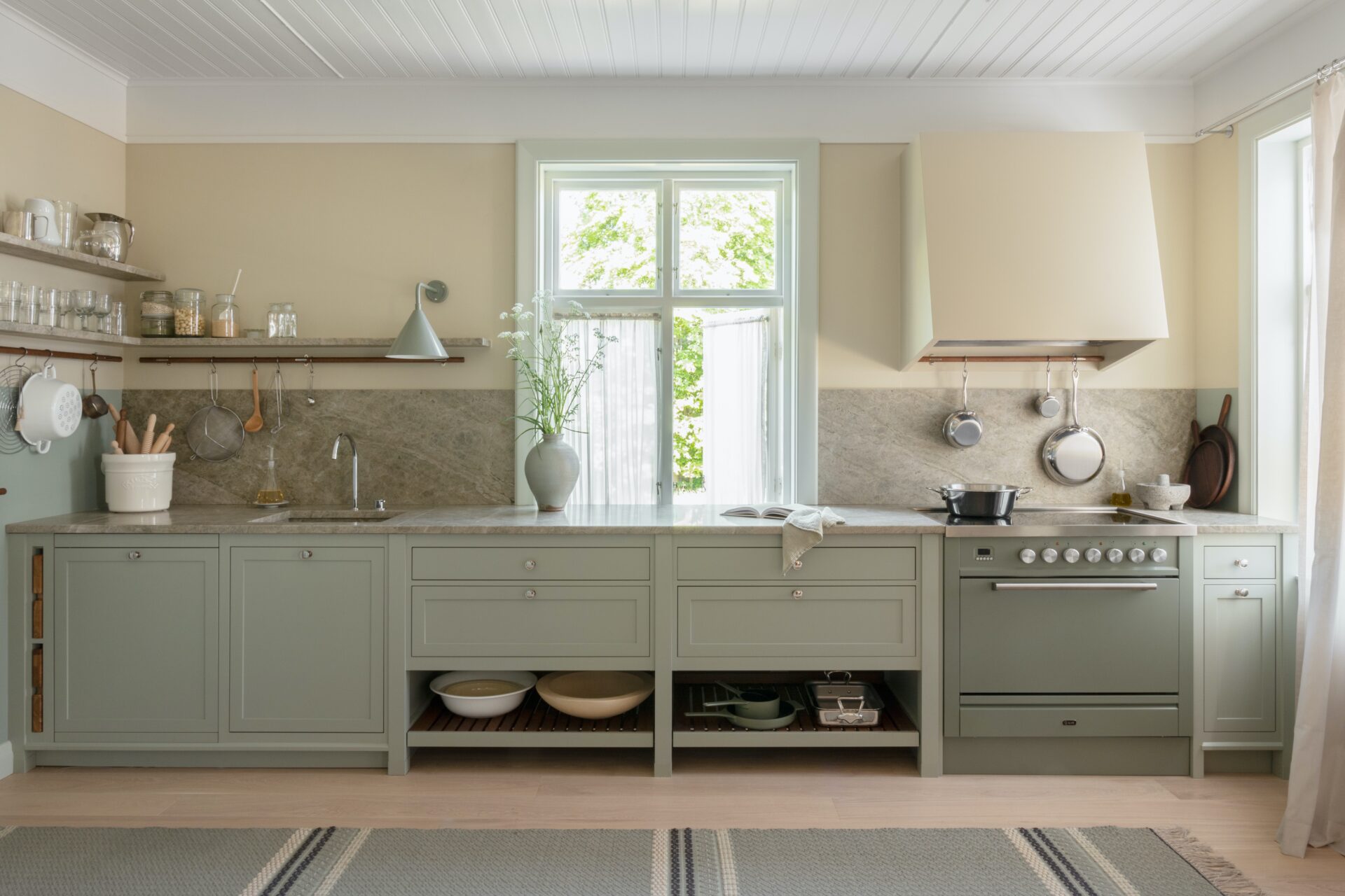 Kök med salviagröna skåp, marmorbänkskiva, spis, ugn, diskho, öppna hyllor och stort fönster för naturligt ljus.