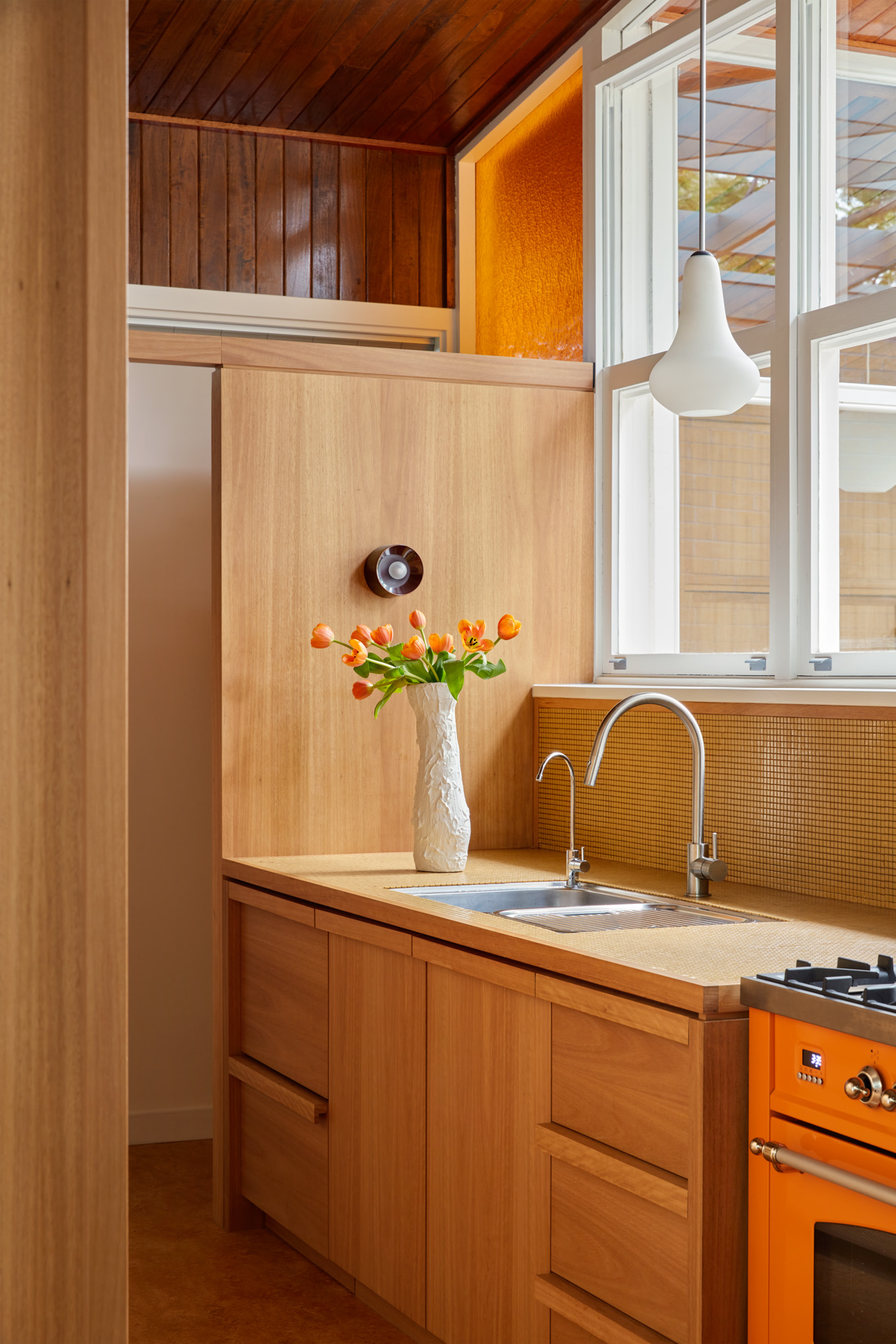 Litet kök med ljusa träskåp, orange spis, vit pendellampa, stort fönster och vas med orange tulpaner vid rostfri diskho.