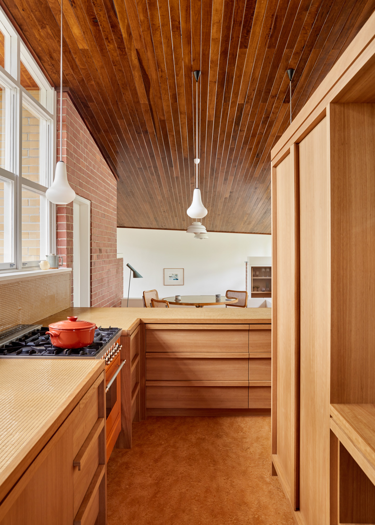 Modernt kök med träskåp, träpanel i taket, terrakottagolv, spis med röd gryta och pendellampor mot ljus vardagsrumsyta.