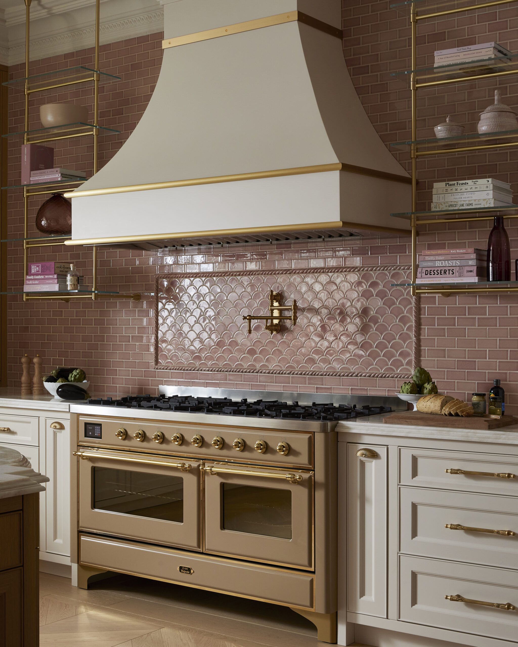 Lyxigt kök med creme- och guldfärgad spis, matchande köksfläkt, rosa kakel, vita skåp samt gyllene hyllor och detaljer.