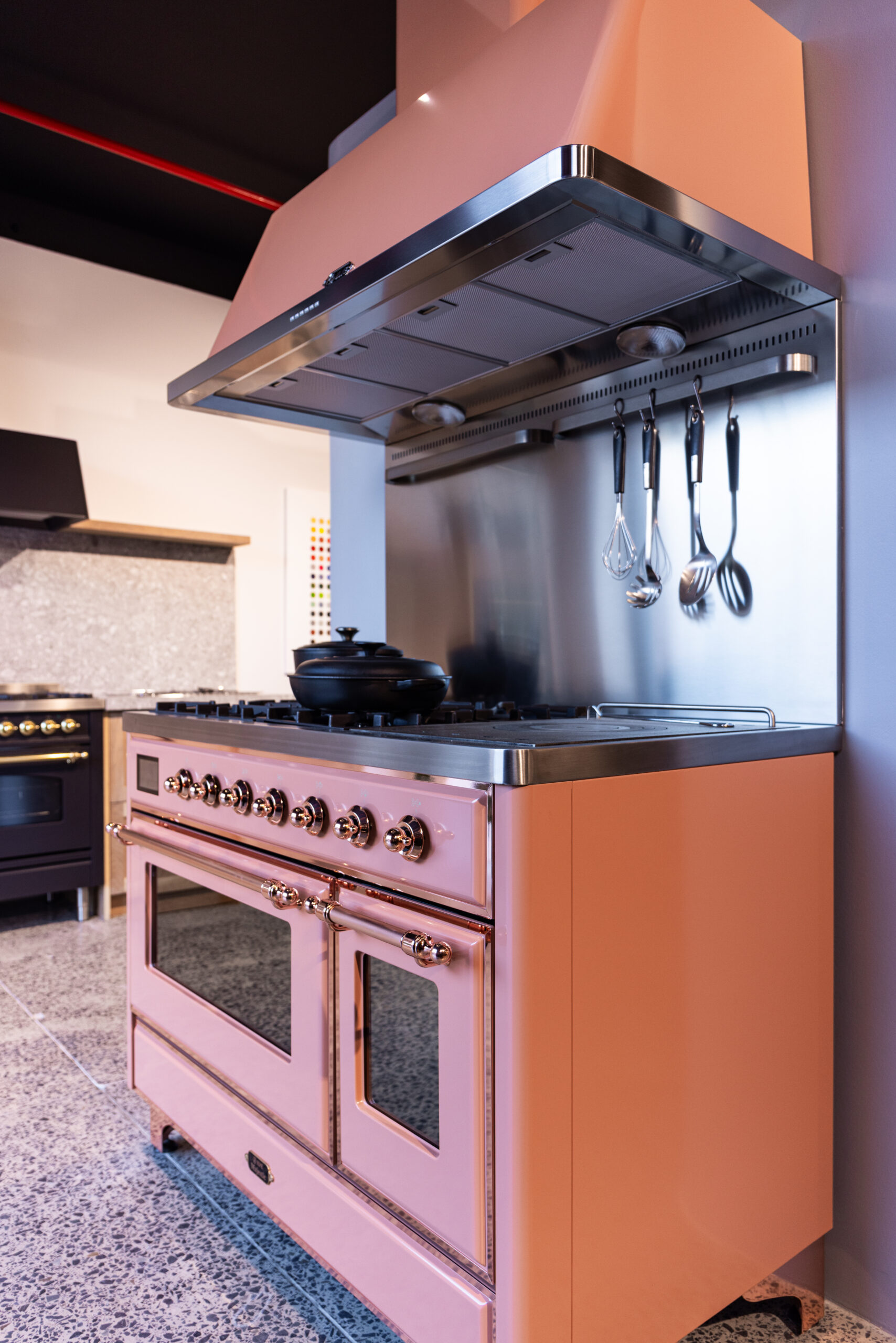 Ett modernt kök med stor rosa och rostfri spis/ugn, hängande köksredskap samt svart gryta på stänkstensgolv; ytterligare svart ugn syns i bakgrunden.