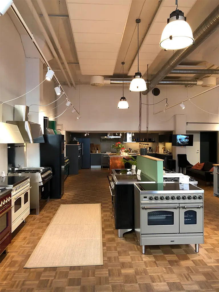 Ett rymligt, modernt showroom med flera spisar, ugnar och andra vitvaror längs väggarna, parkettgolv och pendelbelysning.