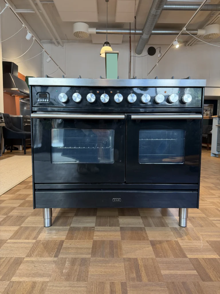 Stor svart dubbellampa med metallreglage och handtag på kromben, placerad på parkettgolv i en modern utställning.