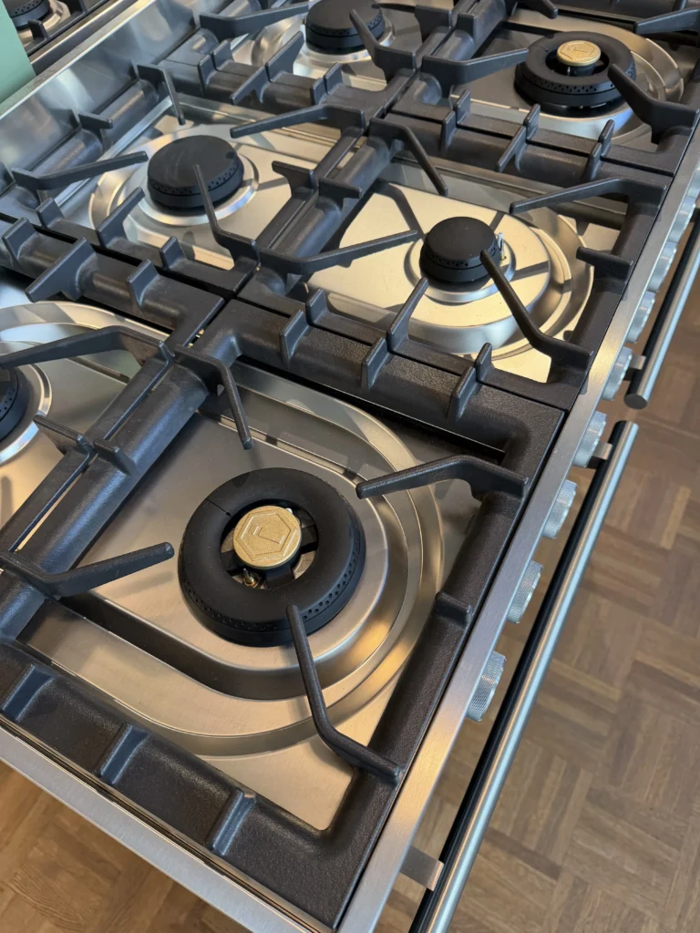 Närbild av en spis i rostfritt stål med fyra brännare, svarta gjutjärnsgaller och metallreglage på trägolv.
